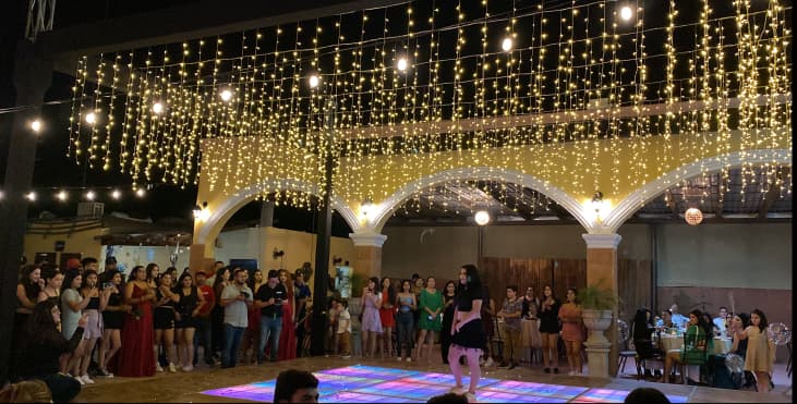 Evento festivo con personas alrededor de una pista de baile iluminada y decorada con luces de hadas. Ambiente cálido y encantador.
