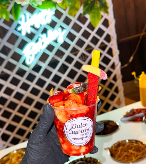 Mano con guante negro sosteniendo un vaso de Dulce Capricho Snacks lleno de frutas y dulces, con fondo de "Happy Birthday" iluminado.