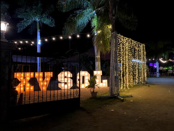 Evento nocturno al aire libre con letras iluminadas "XV SOL" cerca de una puerta, luces colgantes y palmeras en el fondo. Ambiente festivo y decorativo.
