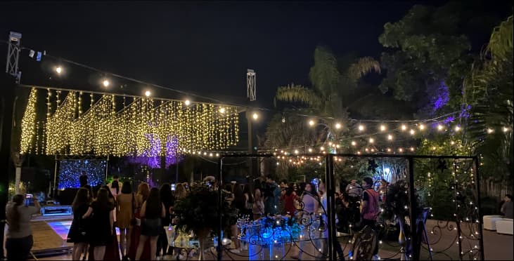Fiesta al aire libre por la noche con luces colgantes, escenario con luces púrpuras y pista de baile iluminada. Ambiente festivo y animado.