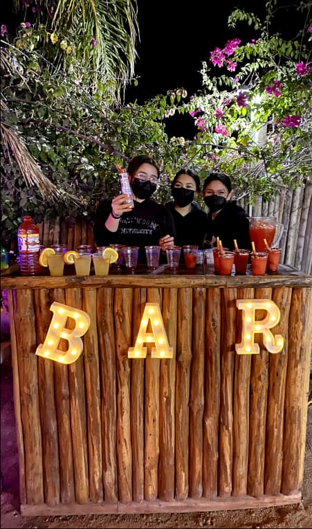 Bar rústico nocturno al aire libre con mostrador de madera y letras iluminadas 'BAR'. Varias bebidas con cócteles y decoración acogedora.