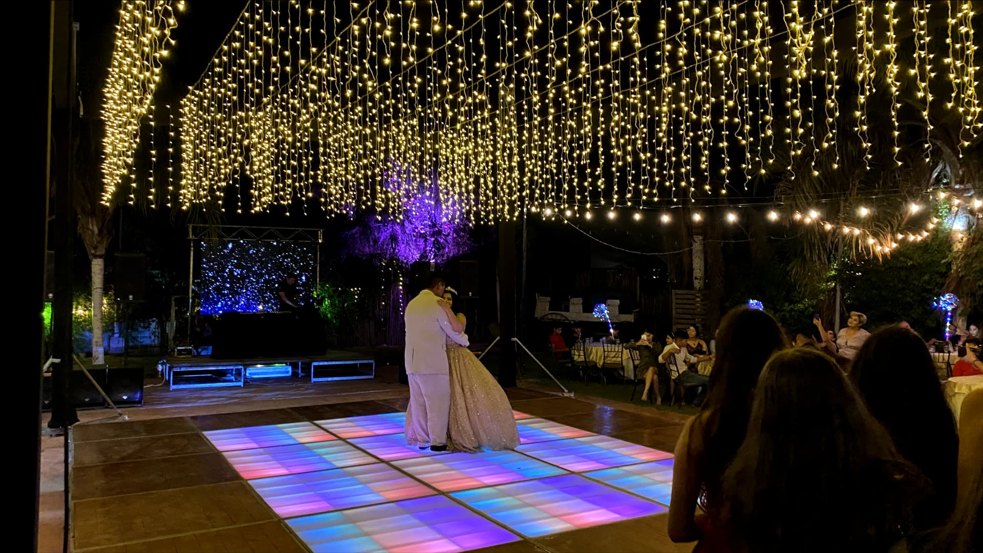 Evento nocturno al aire libre con un baile formal, luces colgantes, pareja bailando y un DJ actuando. Ambiente cálido y elegante.