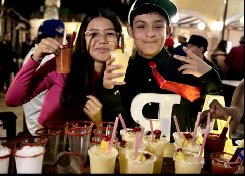 Grupo de personas en una fiesta, con bebidas y decoraciones en la mesa. Ambiente festivo y animado, con personas al fondo.