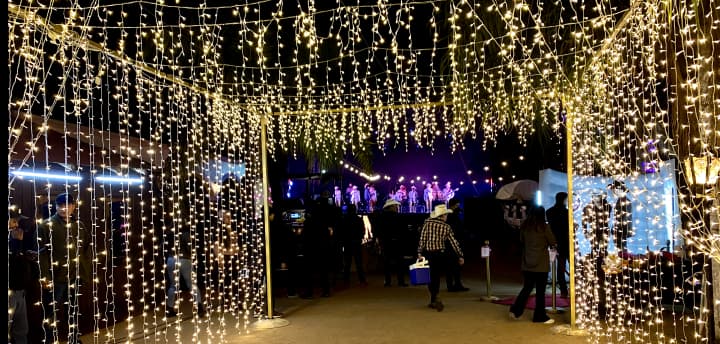 Evento festivo con luces colgantes creando un túnel y un escenario con artistas. Ambiente cálido y acogedor, visualmente atractivo y festivo.
