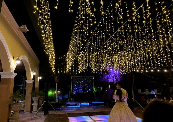 Evento nocturno al aire libre, decorado con luces colgantes, pista de baile iluminada y pareja bailando. Ambiente elegante y festivo.