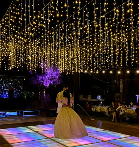 Un evento al aire libre con decoración de luces cálidas y una pista de baile colorida. La atmósfera es festiva y mágica.