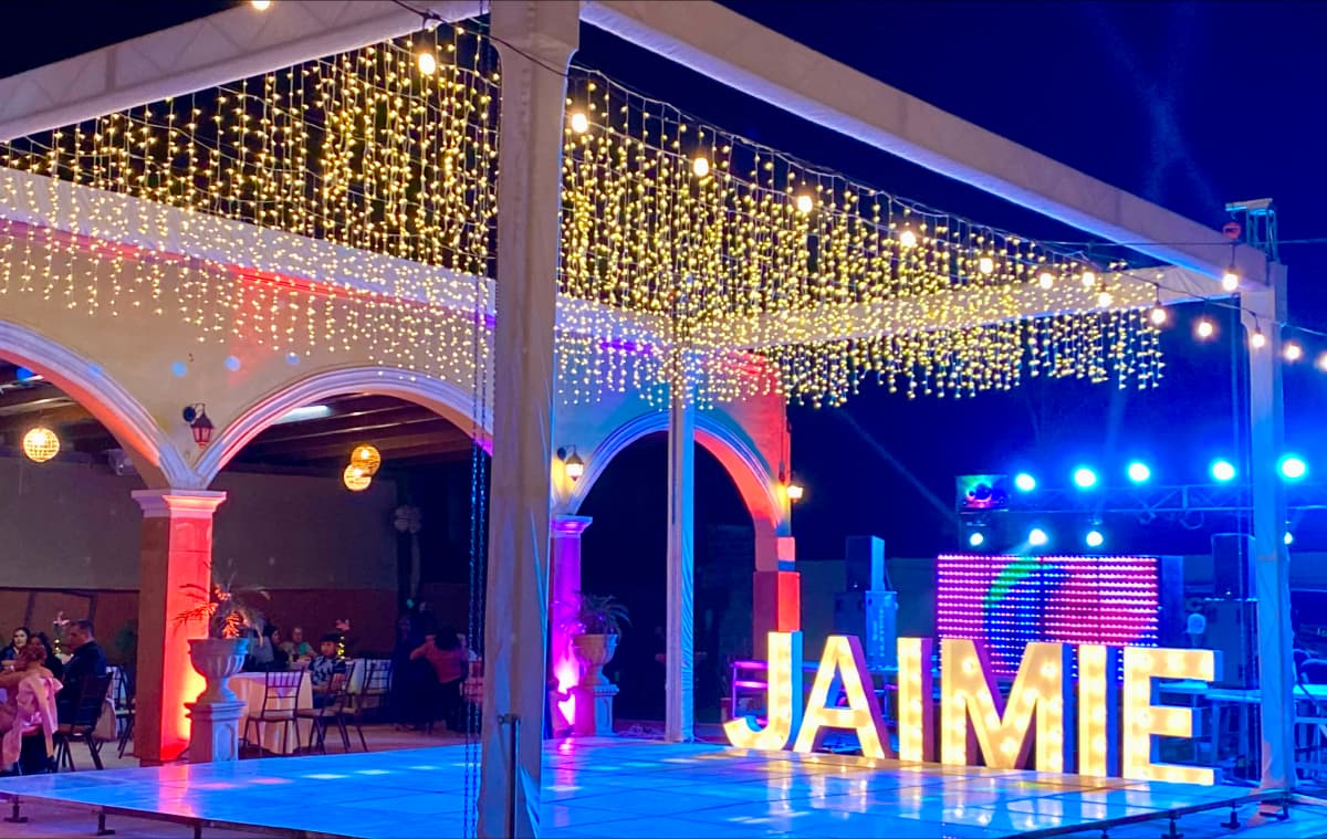 Espacio decorado con luces colgantes, letras iluminadas que deletrean 'JAIMIE', arcos y columnas iluminadas, y mesas con personas sentadas.