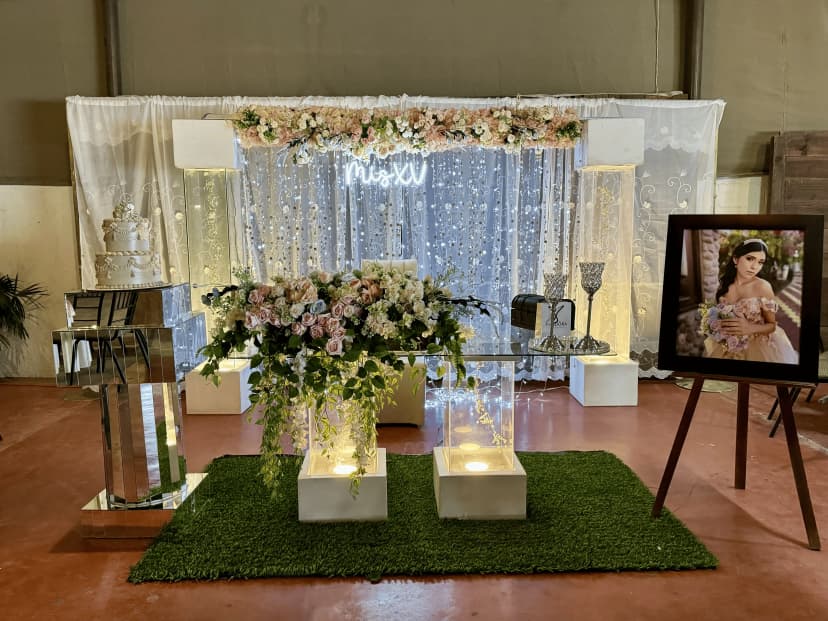 Mesa de honor número 11 con fondo blanco decorado con luces y flores, con letras neón "Mis XV" y un pastel blanco al lado.