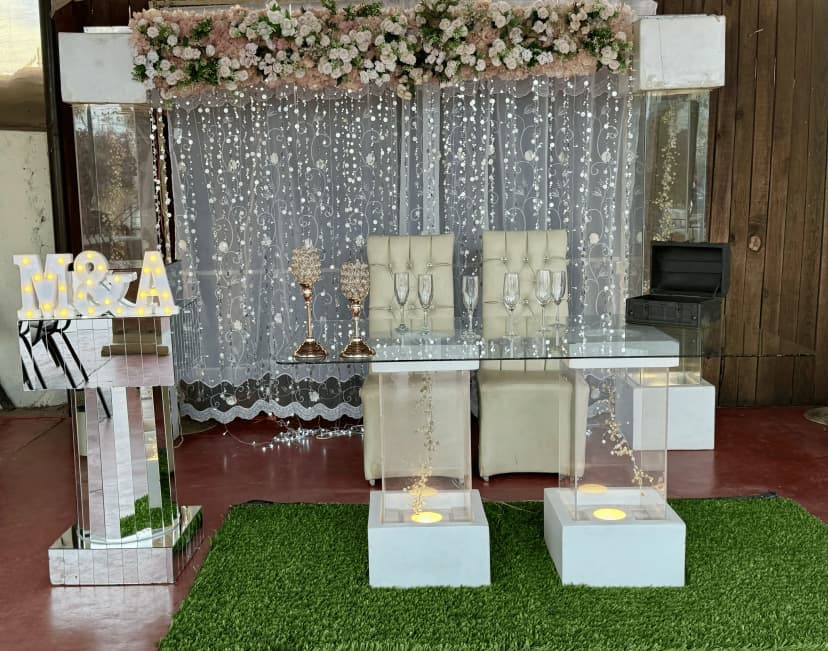 Mesa de honor número 12 con fondo de luces y flores, decorada con copas altas y letras grandes "M&A" en la esquina.