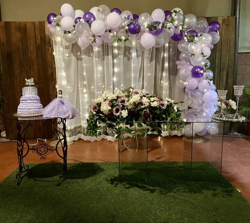 Mesa de honor número 15 decorada con luces y globos en tonos morados, con un elegante arreglo floral y una princesa de juguete.