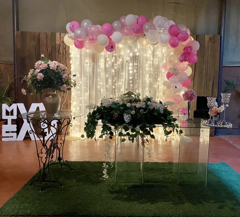 Mesa de honor número 16 con fondo decorado con globos y luces suaves, mesa de cristal con flores en tonos rosas y verdes.