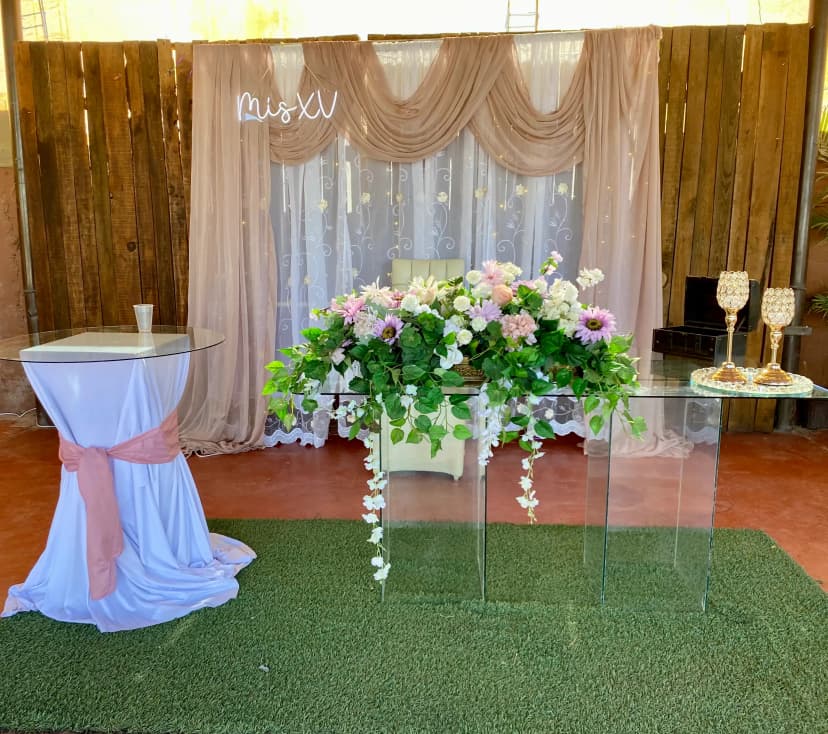 Mesa de honor número 21 con fondo blanco y café, decorada con flores en tonos verdes y morados, con letras neón "Mis XV".