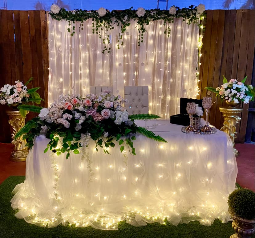 Mesa de honor número 22 con fondo de luces y flores, decorada con un recubrimiento blanco y flores en tonos rosas y verdes.