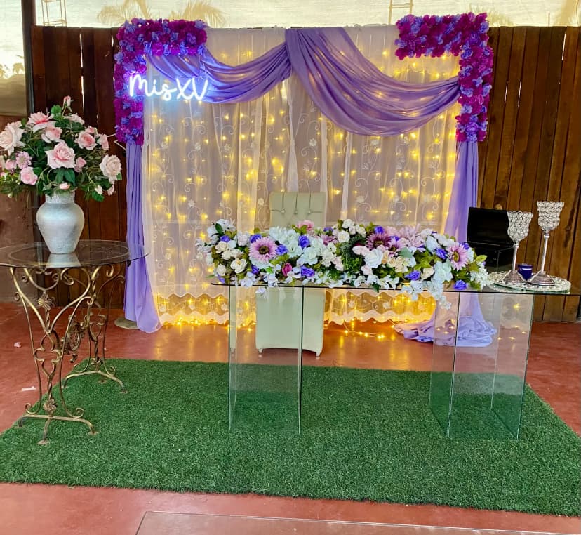 Mesa de honor número 24 con fondo de luces y flores moradas, decorada con flores blancas, moradas y azules en un estilo elegante.