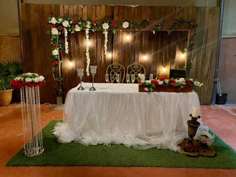 Mesa de honor número 25 con fondo de madera, decorada con luces y flores, con una elegante decoración floral en la mesa blanca.