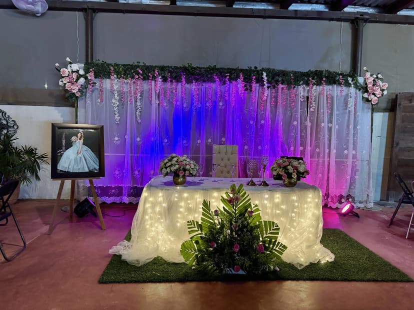 Mesa de honor número 8 con fondo de luces neón azules y rosas, decorada con arreglos florales y un retrato detrás de la mesa blanca.